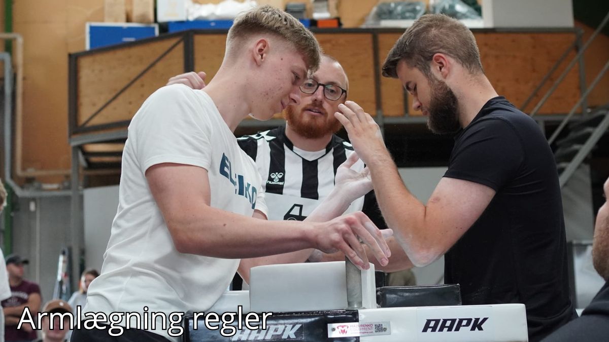 Billedet viser to mænd, der gør sig klar til en armlægningskamp ved et professionelt bord. En dommer i sort-hvid-stribet trøje overvåger dem nøje for at sikre, at de følger reglerne. Baggrunden viser en indendørs hal med tilskuere og andre deltagere, der skaber en konkurrencestemning. Billedetekst: "Armlægningsregler''