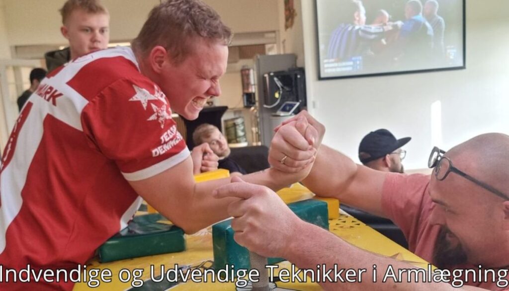 Billedet viser to mænd engageret i en armlægningsduel. Den ene mand, iført en rød trøje med "Team Danmark," udstråler intens koncentration og styrke, mens han kæmper mod en modstander med briller. Baggrunden inkluderer tilskuere og en skærm, der viser en armlægningskamp, hvilket giver en professionel atmosfære. Billedetekst: "Indvendige og Udvendige Teknikker i Armlægning''