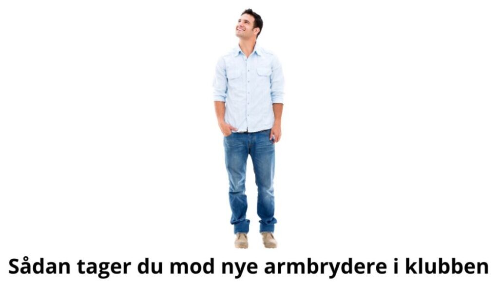 Billedet viser en mand klædt i en lyseblå skjorte og jeans, der står afslappet med hænderne i lommerne og kigger op med et venligt smil. Baggrunden er helt hvid, hvilket giver et enkelt og imødekommende udtryk. Billedetekst: "Sådan tager du mod nye armbrydere i klubben."