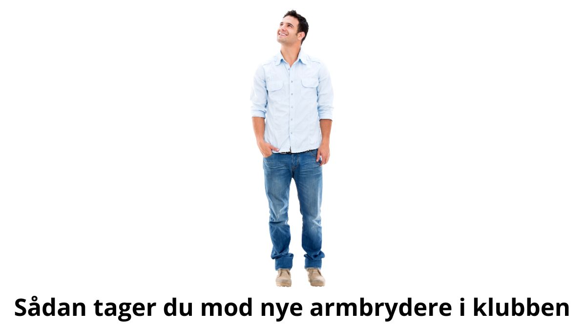 Billedet viser en mand klædt i en lyseblå skjorte og jeans, der står afslappet med hænderne i lommerne og kigger op med et venligt smil. Baggrunden er helt hvid, hvilket giver et enkelt og imødekommende udtryk. Billedetekst: "Sådan tager du mod nye armbrydere i klubben."