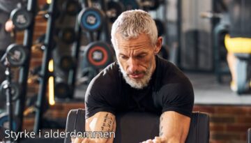 Billedet viser en ældre mand med gråt hår og skæg, der træner koncentreret i et fitnesscenter. Han udfører en øvelse for armstyrke og ser fokuseret og målrettet ud. Bag ham ses vægte og træningsudstyr, som understreger miljøet. Billedetekst: "Styrke i alderdommen''