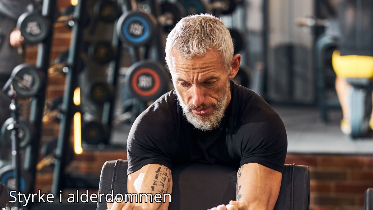 Billedet viser en ældre mand med gråt hår og skæg, der træner koncentreret i et fitnesscenter. Han udfører en øvelse for armstyrke og ser fokuseret og målrettet ud. Bag ham ses vægte og træningsudstyr, som understreger miljøet. Billedetekst: "Styrke i alderdommen''