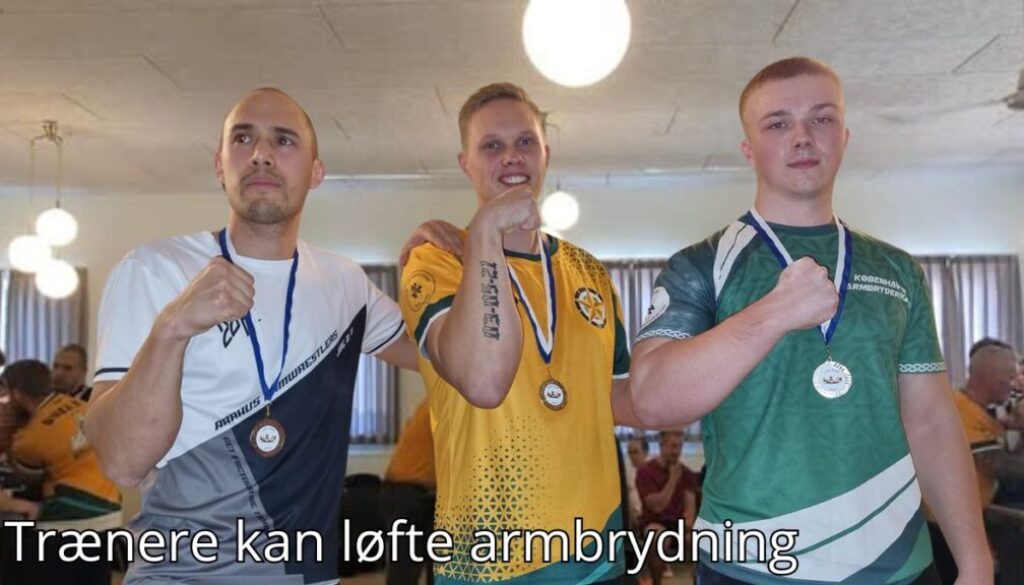 Billedet viser tre mænd iført holdspecifikke trøjer, der stolt fremviser deres medaljer og poserer med hænderne knyttet i en sejrspositur. Baggrunden viser en indendørs hal med andre deltagere og tilskuere, hvilket skaber en stemning af konkurrence og fællesskab. Billedetekst: "Trænere kan løfte armbrydning''