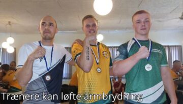 Billedet viser tre mænd iført holdspecifikke trøjer, der stolt fremviser deres medaljer og poserer med hænderne knyttet i en sejrspositur. Baggrunden viser en indendørs hal med andre deltagere og tilskuere, hvilket skaber en stemning af konkurrence og fællesskab. Billedetekst: "Trænere kan løfte armbrydning''
